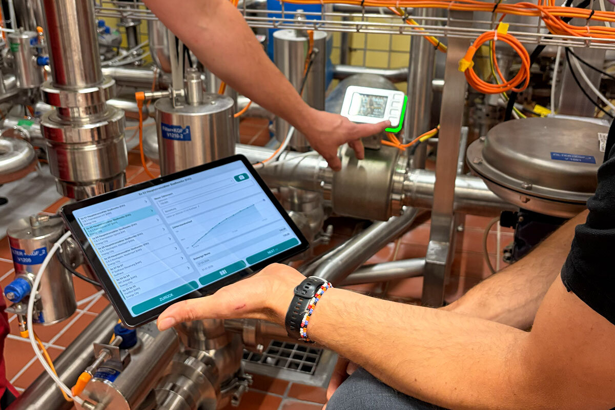 Digitales Energiemanagement in der Brauerei: Eine Person prüft Messwerte an Edelstahlrohren mit Messgerät und Tablet.