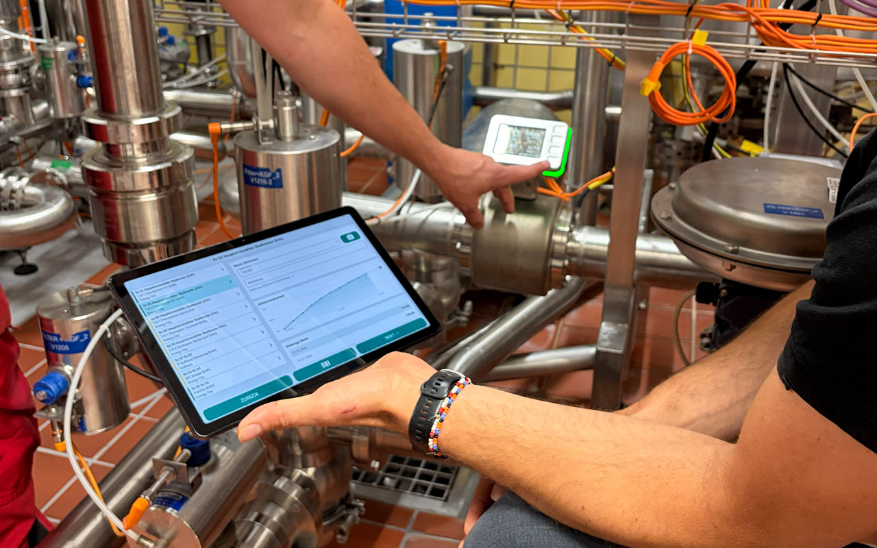 Digitales Energiemanagement in der Brauerei: Eine Person pr&uuml;ft Messwerte an Edelstahlrohren mit Messger&auml;t und Tablet.